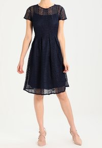 Laivastonsininen pitsimekko, jossa on lyhyet hihat, istuva yläosa ja levenevä hame. Sisältää monimutkaisia leikattuja kuvioita ja polvipituista muotoilua.