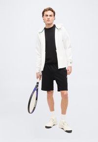 Νέος άνδρας με λευκό σακάκι, μαύρο πουκάμισο και σορτς, κρατώντας ρακέτα τένις σε χαλαρή στάση σε απλό φόντο.