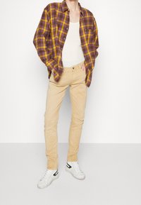 Camisa de cuadros marrón y amarillo, camiseta blanca sin mangas, pantalones beige claros y zapatillas blancas con detalles en negro; ajuste casual con un cuello relajado.