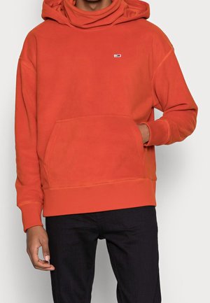 Person, die einen orangefarbenen Kapuzenpullover mit Fronttasche und kleinem Logo trägt, kombiniert mit schwarzen Hosen, vor einem einfarbigen hellen Hintergrund.