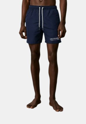 Modello maschile snello che indossa pantaloncini da bagno blu navy con cordoncino bianco e logo Trussardi sulla gamba sinistra, in piedi a piedi nudi su uno sfondo uniforme.