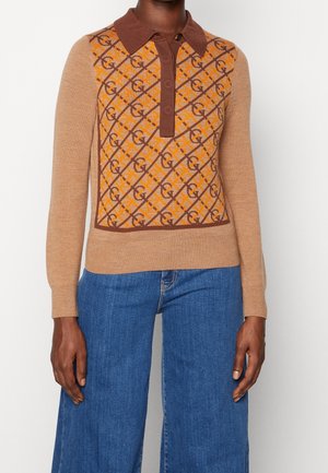 Beżowy sweter z długim rękawem z brązowym kołnierzem i pomarańczowo-brązowym geometrycznym wzorem z przodu, noszony z niebieskimi jeansami o szerokich nogawkach.