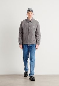 Geacă din fleece gri cu nasturi și logo HUGO, asortată cu blugi albaștri și pantofi negri. Modelul poartă o căciulă gri cu literele HUGO.