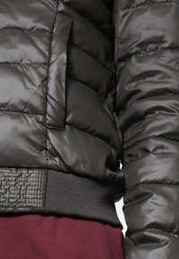 Svart puffjacka med en glansig finish, quiltad textur och ribbade ärmslut. Inkluderar en sidoficka med ett subtilt mönster.