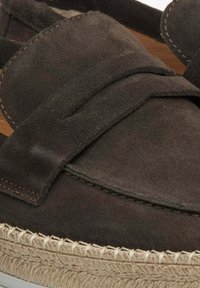 Bruine suede instapper met een brede band over de voorkant, voorzien van een gevlochte jute zool en witte stiksels langs de rand.