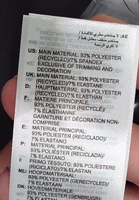 Kleidehoolduse silt erinevates keeltes, kus on kirjas 93% taaskasutatud polüester ja 7% spandeks/elastaan; trükitud tekstuursele kangale.