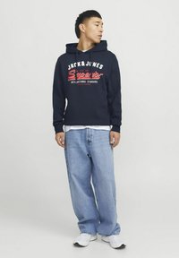 Marineblauer Hoodie mit Grafik "JACK & JONES Superior" in Rot und Weiß. Kombiniert mit hellblauen weiten Jeans und weißen Sneakern.