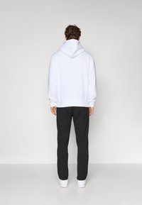 Weißer Kapuzenpullover mit glatter Textur, gerippten Bündchen und Saum. Kombiniert mit dunklen Hosen und weißen Sneakers, von hinten betrachtet.