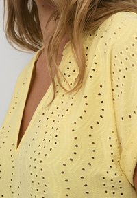 Keltainen puuvillapaitapusero, jossa on V-pääntie, brodeerattu kuvio leikkauksilla ja lyhyet hihat.