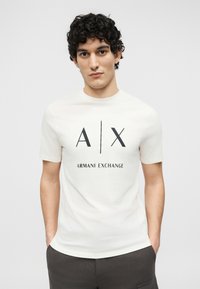 Νεαρός με μπούκλες που φοράει λευκό T-shirt Armani Exchange με μαύρο λογότυπο, τα χέρια στις τσέπες, μπροστά σε απλό ανοιχτόχρωμο φόντο.