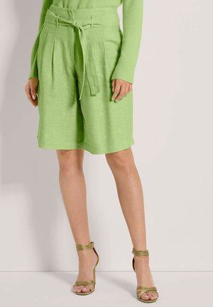 Hellgrüne Leinen-Shorts mit Falten, einem gebundenen Taillenbund und lockerem Schnitt. Kombiniert mit olivgrünen Riemchen-Absatzschuhen.