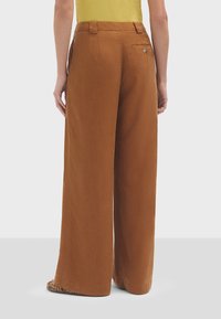 Pantalons larges marron en tissu doux. Présente des poches arrière et des coutures surpiquées. Associés à un haut jaune, présentés de dos.