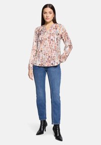 Durchsichtige Bluse mit langen Ärmeln und einem mehrfarbigen runden Muster, ausgestattet mit einem V-Ausschnitt und gerafften Manschetten, kombiniert mit blauen Jeans und schwarzen Knöchelschuhen.