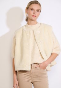 Faux fur gilet in crème met een ronde halslijn, met knopen aan de voorkant en zonder mouwen, gecombineerd met een gestructureerde beige top en bijpassende broek.