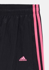 Črne športne hlače z roza stranskimi črtami, elastičnim pasom in teksturirano tkanino. Vključujejo majhen roza Adidas logotip.