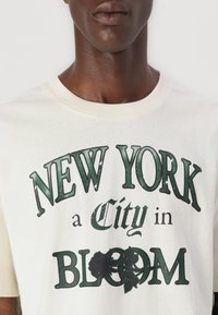 Cremefarvet bomulds t-shirt med grøn grafisk tekst "NEW YORK a City in BLOOM," med blomsterdetaljer og en blød, afslappet pasform.