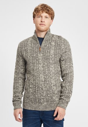 Suéter gris de punto con cuello alto y cierre de media cremallera. Presenta patrones texturizados y puños acanalados. Se lleva con pantalones vaqueros de color azul oscuro.