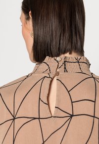 Blouse beige avec des motifs géométriques noirs, col montant avec des détails froncés, et fermeture à deux boutons à l'arrière. Texture de tissu douce.