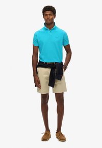 Camisa de polo turquesa, de manga corta, con cuello. Pantalones cortos beige con un cinturón, suéter oscuro atado en la cintura y zapatos marrones de cordones.