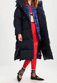 Dlouhý námořnický puffer kabát s kapucí, vybavený velkými prošívanými sekcemi, stříbrnými zipy, přes červený outfit s síťovými akcenty a kostičkovanou košilí.