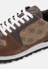 Braune Sneaker mit einem Obermaterial aus Wildleder und gemustertem Stoff, weißen Schnürsenkeln, Gummisohle und einem kleinen metallischen Logo-Akzent an der Zunge.