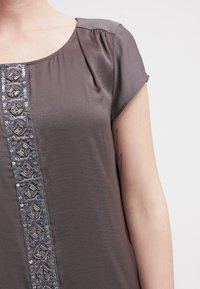 Top gris con mangas cortas, que presenta un panel decorativo con lentejuelas y acentos geométricos en el centro. Tejido suave con un ligero brillo.