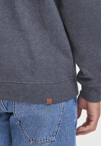 Sweatshirt en coton gris avec des poignets et un ourlet côtelés, arborant une petite étiquette marron en bas. Assorti à un jean en denim bleu délavé.