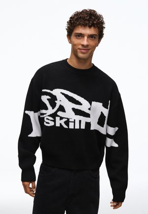 Jeune homme aux cheveux bouclés portant un pull noir avec un grand texte abstrait blanc, debout devant un fond blanc uni.