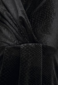 Tissu en velours noir avec un motif subtil de peau de serpent, rassemblé et enveloppé d'une ceinture texturée assortie.
