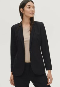 Zwarte op maat gemaakte blazer met een enkele knoopsluiting, notch-revers en twee zijzakken, gemaakt van gladde stof. Draagt over een lichtbeige top.