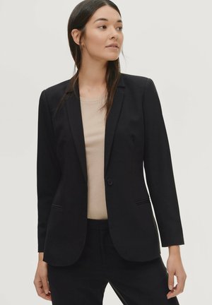 Zwarte op maat gemaakte blazer met een enkele knoopsluiting, notch-revers en twee zijzakken, gemaakt van gladde stof. Draagt over een lichtbeige top.