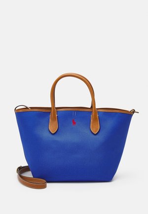 Niebieska torba typu tote z brązowymi skórzanymi uchwytami i wykończeniem, małym czerwonym logo umieszczonym centralnie między uchwytami oraz odpinanym paskiem na ramię na białym tle.