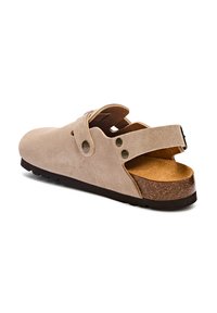 Beige suède klomp met verstelbare band, kurken voetbed, rubberen zool en open achterkant. Kenmerkt zich door twee decoratieve metalen klinknagels en een gestructureerde bovenkant.