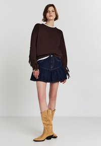 Donna in piedi che indossa un maglione fringed marrone, una gonna di denim blu e stivali da cowboy beige su uno sfondo chiaro e uniforme.