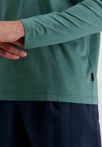 Mann, der den Ärmel eines grünen Langarmhemds anpasst, trägt dunkelblaue Hosen vor einem schlichten, hellen Hintergrund.