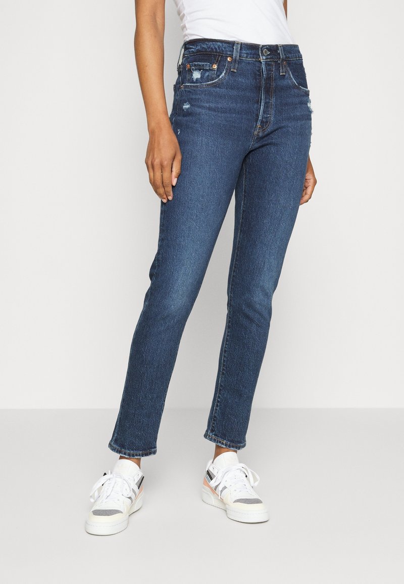 Personne portant un jean slim bleu foncé légèrement déchiré, des baskets blanches et un top sans manches blanc rentré, debout devant un fond uni.