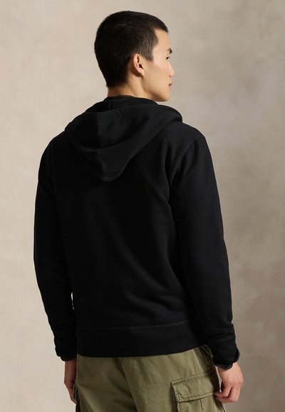 Moški v prečni pozi, oblečen v črno jopa s kapuco in olivno zelene hlače cargo na enobarvnem bež ozadju.