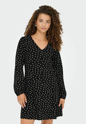 Jeune femme aux cheveux bouclés portant une robe noire à manches longues avec un petit motif floral blanc, se tenant devant un fond clair.