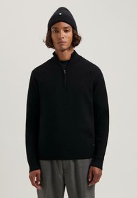 Pulover negru cu guler tip fermoar și manșete ribbed, fabricat dintr-un material tricotat texturat. Asortat cu o căciulă neagră cu un accent verde.