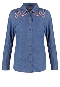 Chemise en denim bleu à manches longues avec boutons sur le devant et broderie géométrique rose et noire au niveau de la poitrine et des épaules.