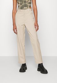 Pantalons beige ajustés avec une coupe droite, en coton, et une fermeture à bouton. Associés à des bottes noires à semelle épaisse.
