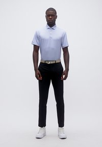 Άνδρας όρθιος, φορώντας γαλάζιο πόλο, μαύρο παντελόνι, μπεζ ζώνη και λευκά αθλητικά παπούτσια σε απλό λευκό φόντο.