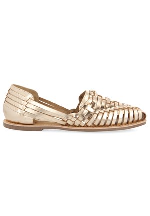 Metalen gouden platte schoenen met een geweven ontwerp, open zijkanten en een platte zool. Het bovenwerk heeft een gladde textuur met een subtiele glans.
