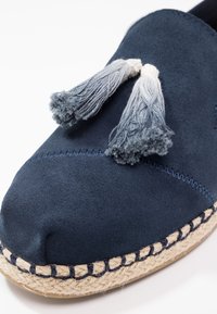 Pantofola in camoscio blu navy con suola intrecciata in stile espadrilla e frange decorative grigie. Presenta cuciture tonali lungo la parte superiore.