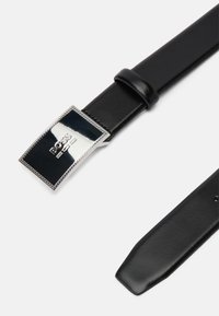 Μαύρη δερμάτινη ζώνη με ορθογώνια, γυαλιστερή ασημένια αγκράφα που φέρει γραμμένο λογότυπο και υφασμάτινο περίγραμμα. Λείο φινίρισμα, κομψός σχεδιασμός.