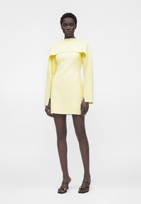 Femme portant une mini-robe jaune pâle à manches longues avec des épaules larges et des sandales noires à talons, debout devant un fond blanc.