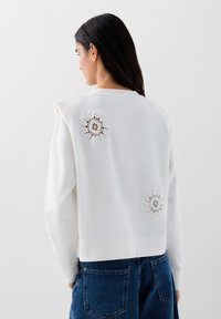 Felpa bianca con ricami floreali sulla schiena, caratterizzata da una vestibilità ampia e una texture morbida, abbinata a jeans in denim blu.