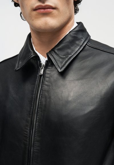 Κοντινό πλάνο ατόμου που φοράει μαύρο δερμάτινο μπουφάν με φερμουάρ μπροστά και μυτερό γιακά πάνω από λευκό πουκάμισο.