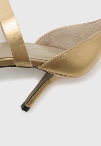 Gold metallic high-heeled shoe with cross straps and a pointed toe. The textured upper contrasts with the smooth insole and slim heel.
