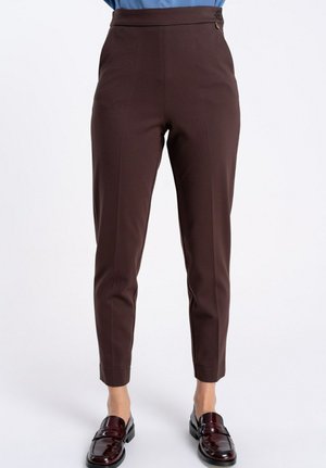 Brązowe, dopasowane do ciała spodnie sięgające kostek, noszone z ciemnoczerwonymi skórzanymi mokasynami przez stojącą osobę na jednolitym tle.